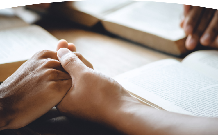 Two hands clasped in prayer beside an open Bible on a wooden table
