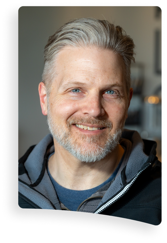 Smiling man with gray hair and beard in a gray hoodie, standing indoors against a softly blurred background