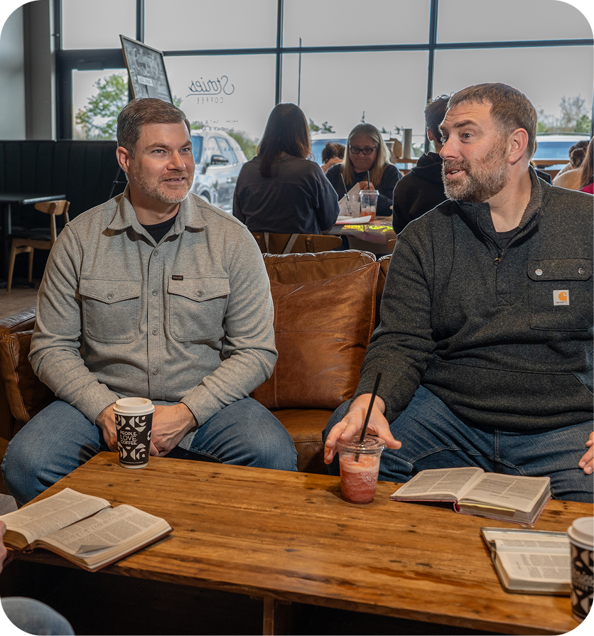 Two people sit at a wooden table in a café, talking with drinks and notebooks open.