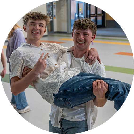Two smiling people in a bright indoor hallway, one carrying the other in their arms.