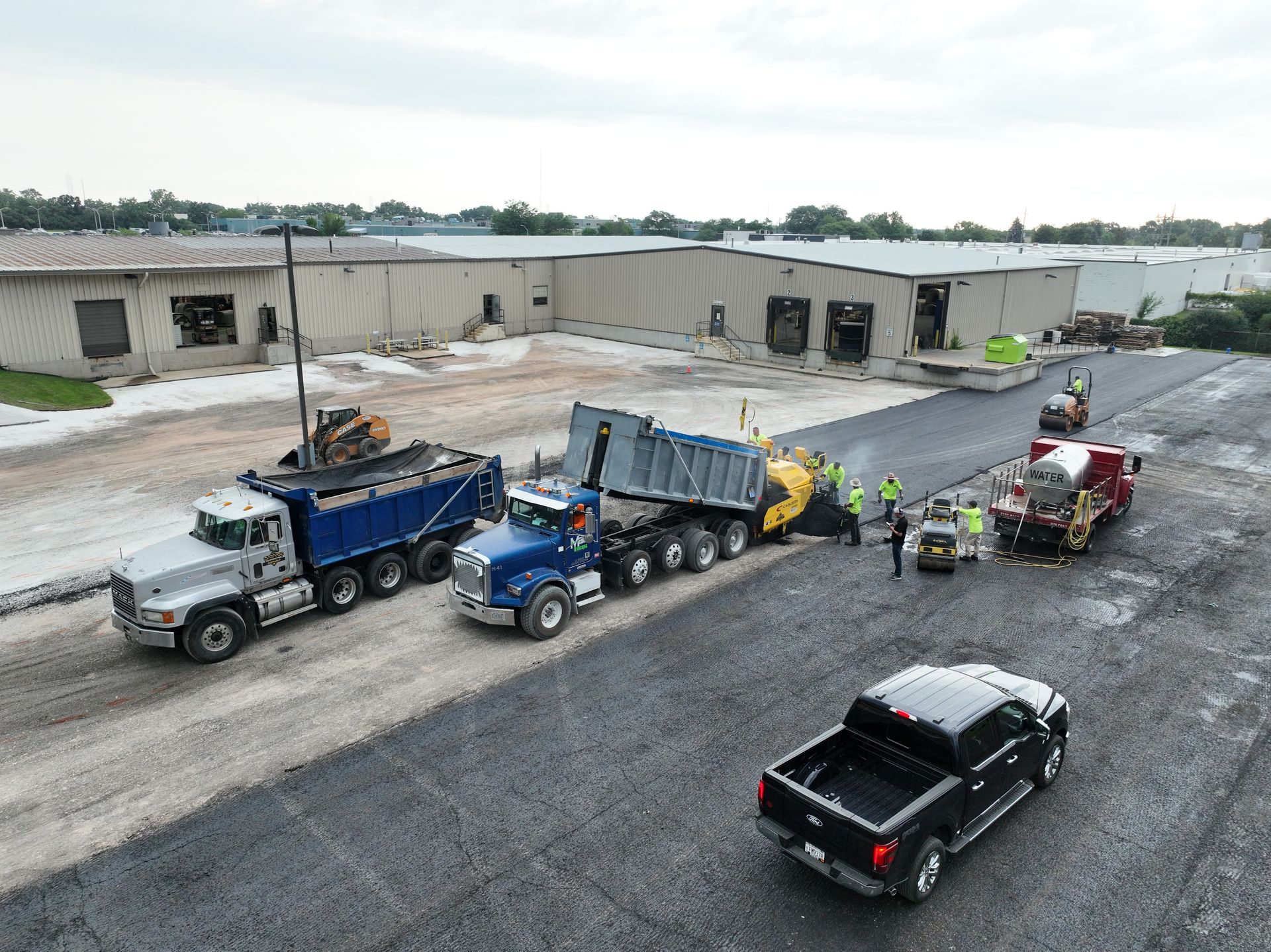 Construction trucks lined up in a gravel lot beside a warehouse, with workers nearby.