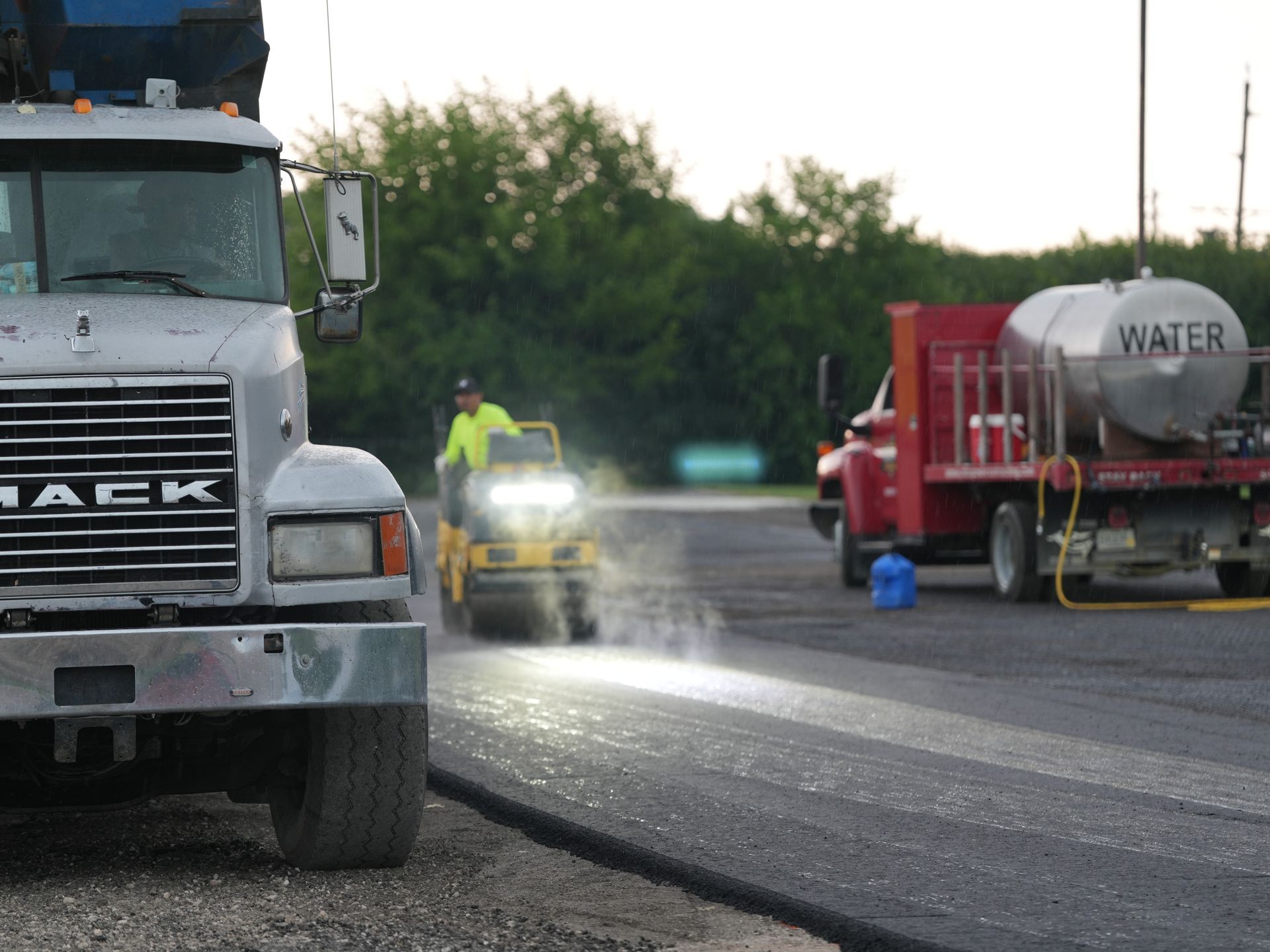 Road work scene with a white truck, worker, and water tanker spraying a wet road at dusk