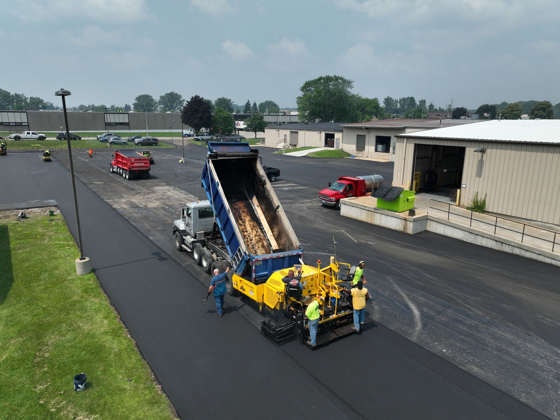 Construction crew paving a road beside a dump truck and red vehicles on an industrial lot