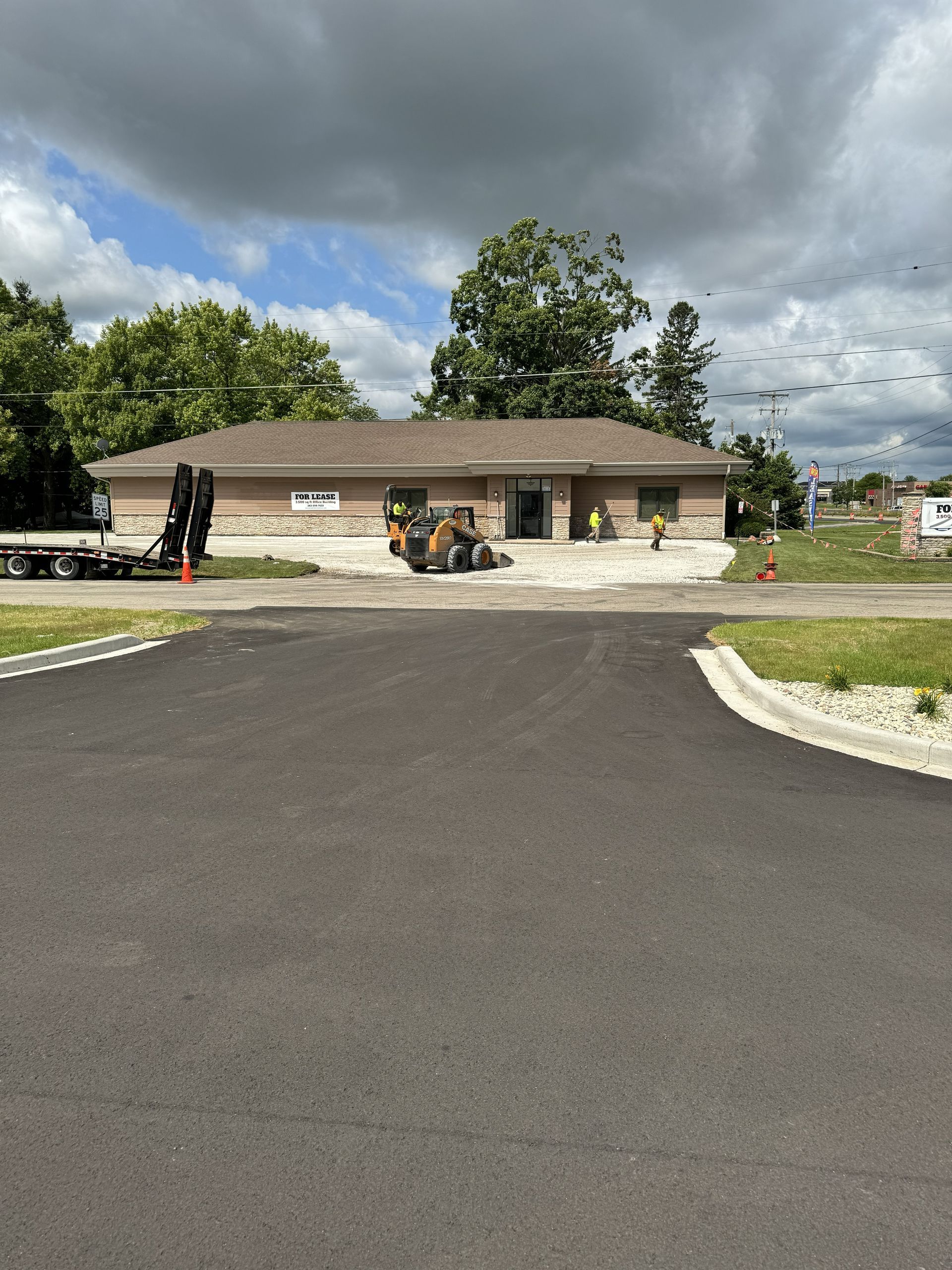 Newly paved road leading to a low brick building under cloudy skies, with landscaping and construction equipment.