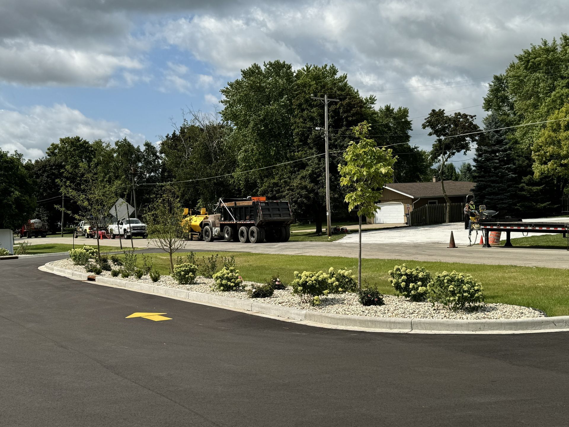 New asphalt road around a landscaped roundabout with shrubs, trees, and a truck in the background