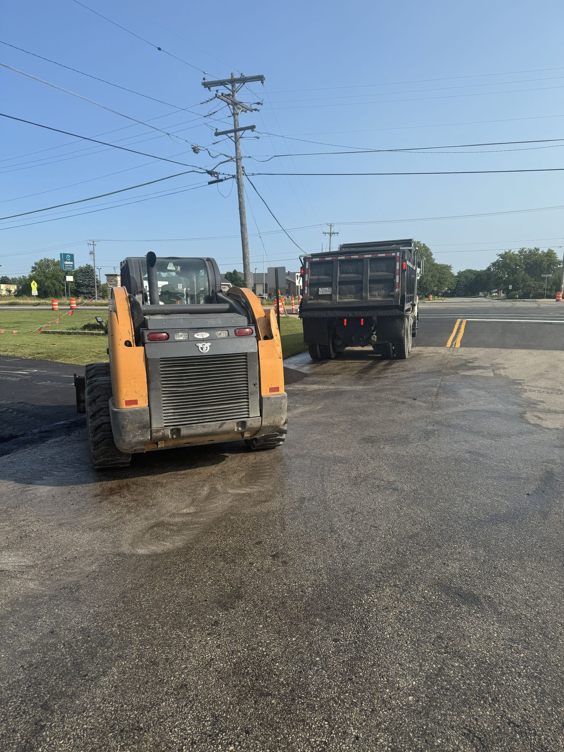 Two construction vehicles on gravel near power lines, one orange skid steer facing a dump truck.