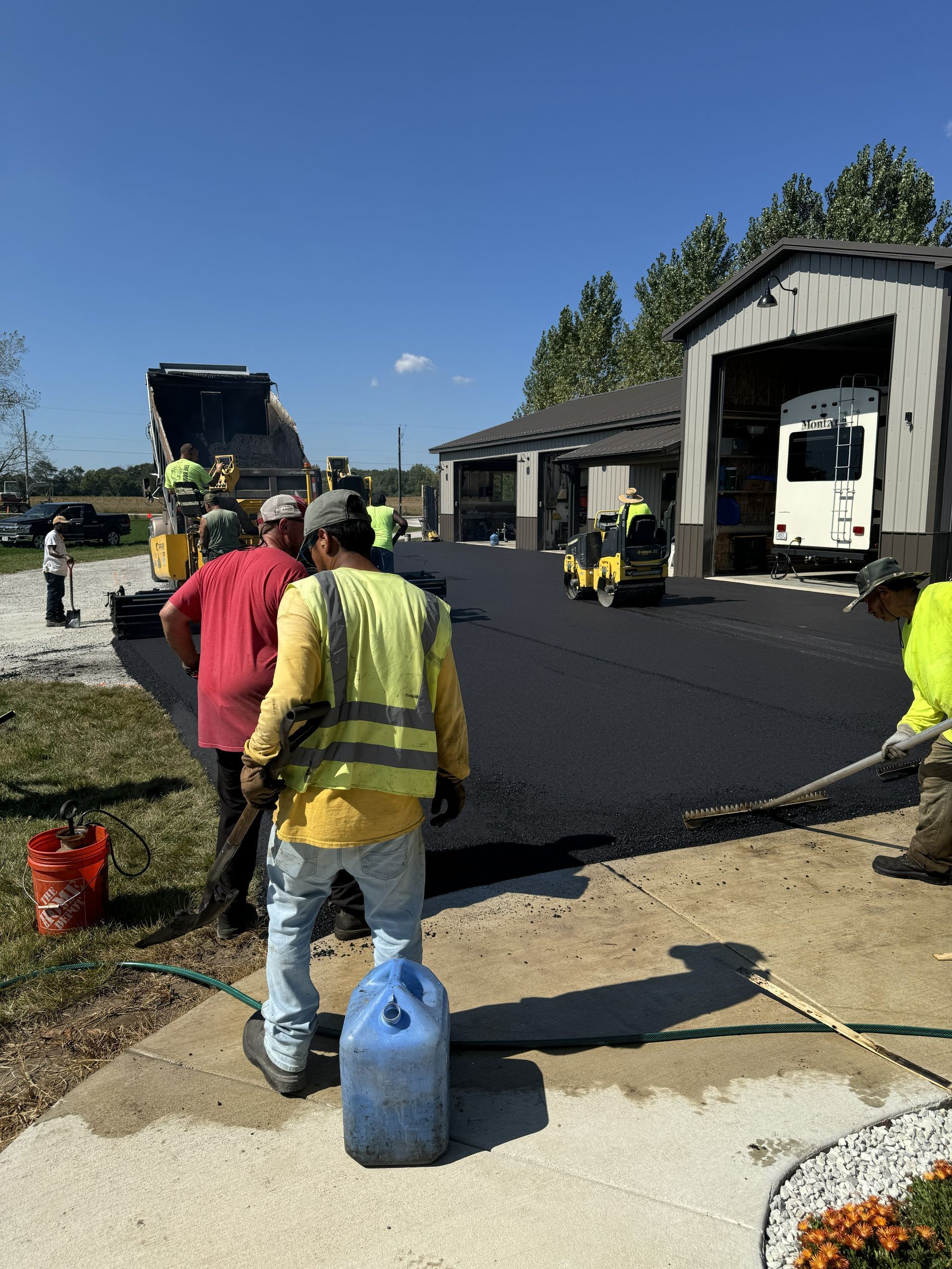 Road crew paving driveway outside garages, with workers and equipment under a clear blue sky