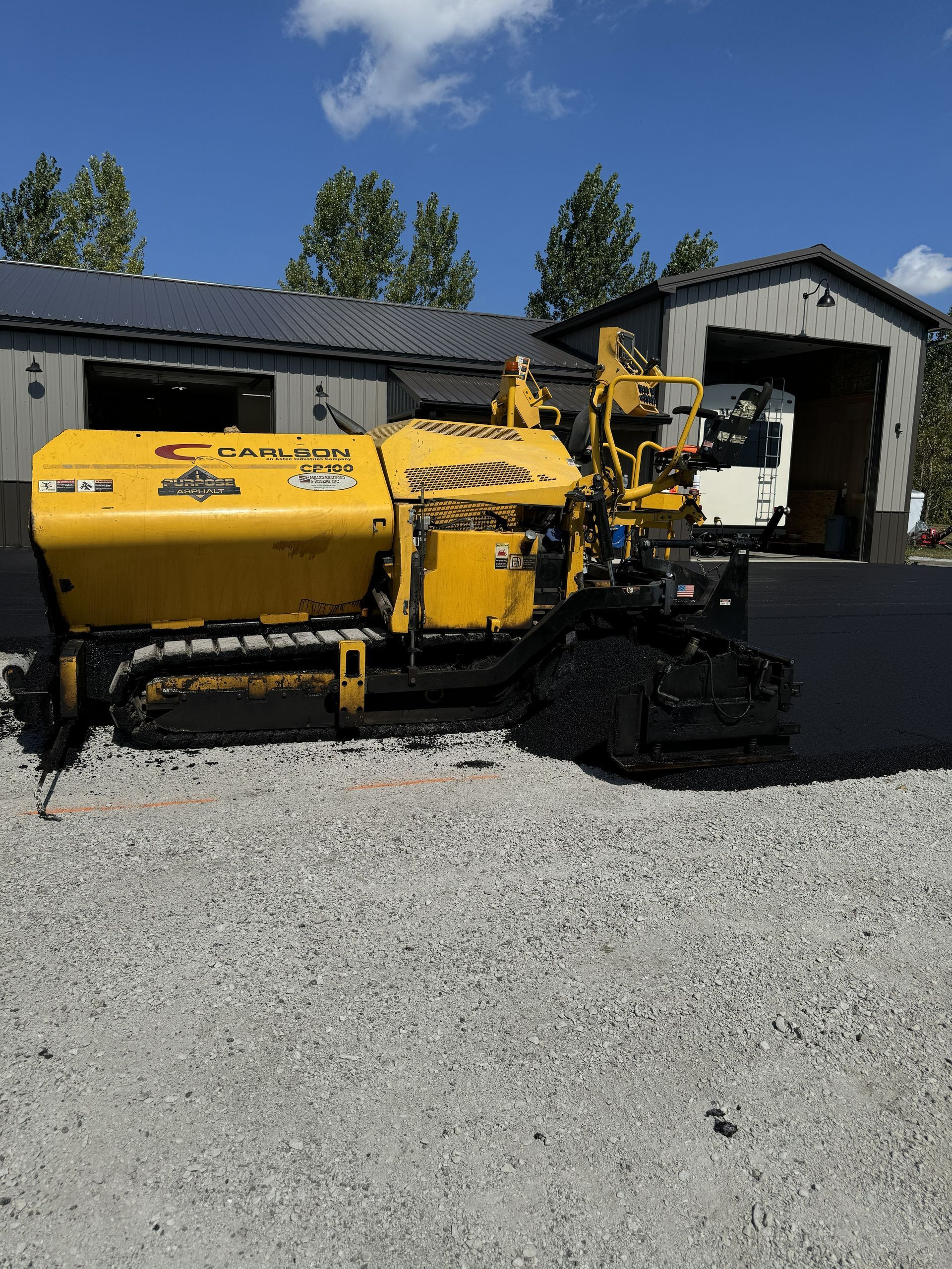 Yellow road construction machine on gravel beside a garage and shed under a blue sky.
