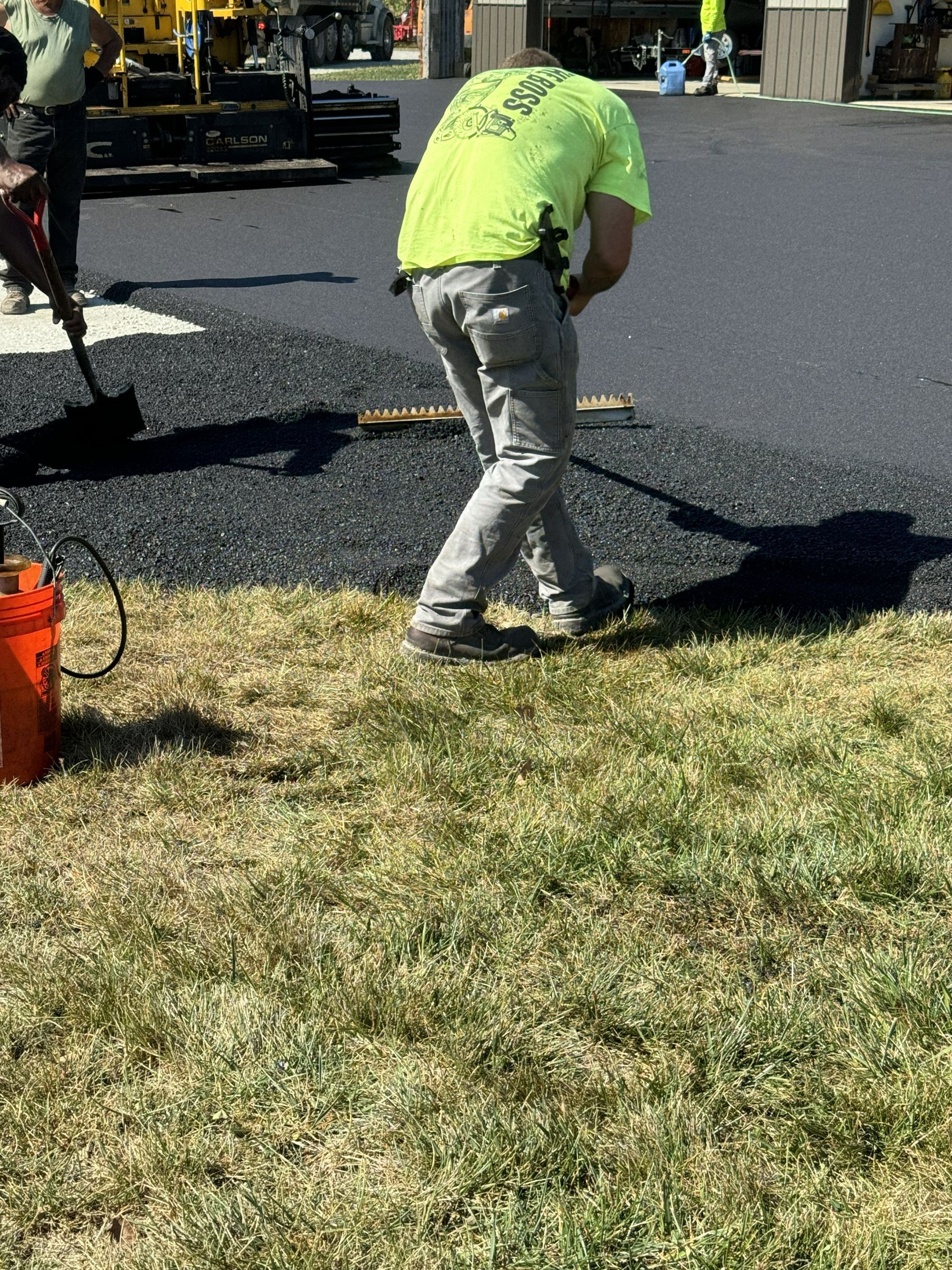 Worker in a neon shirt spreading black asphalt on a road beside a grassy curb.
