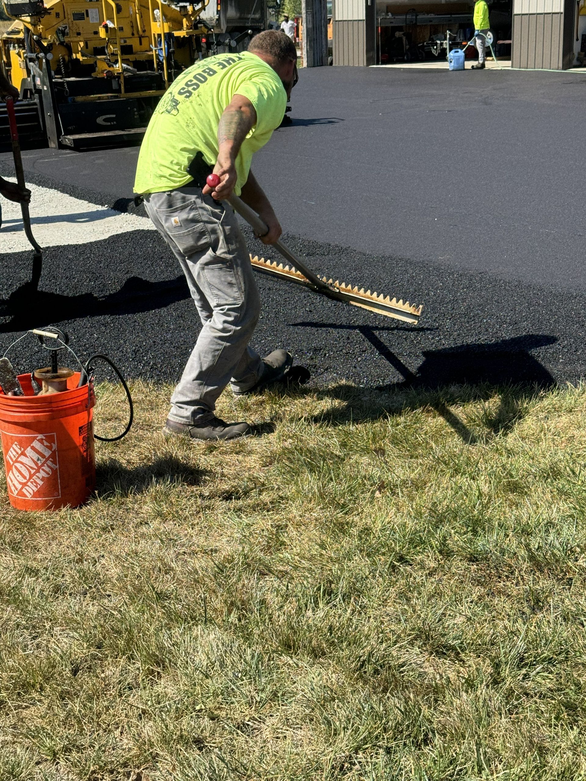 Worker in neon green shirt raking fresh black asphalt beside a grassy edge