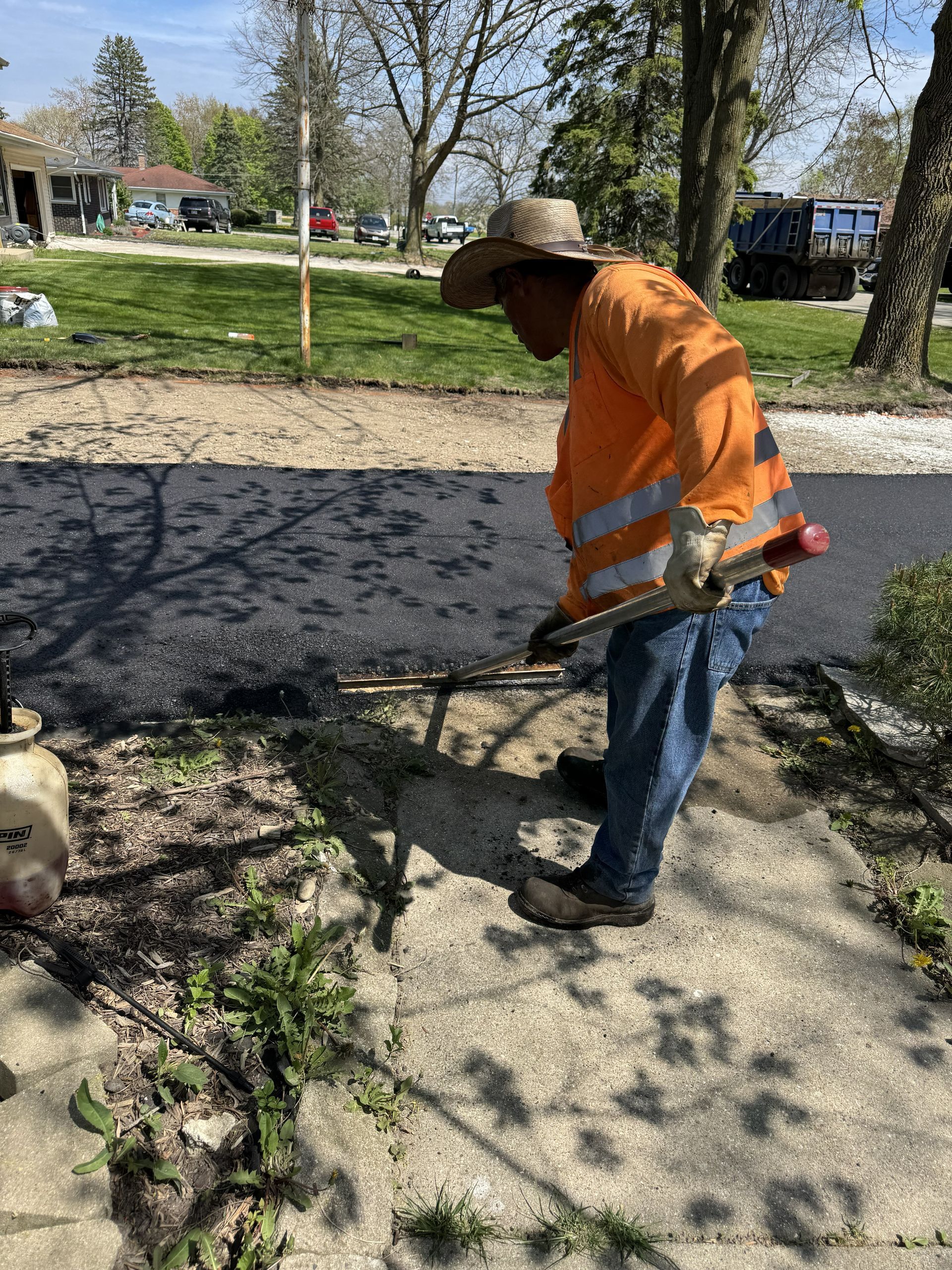 Worker in orange safety vest using a string trimmer beside a sidewalk in a sunny yard
