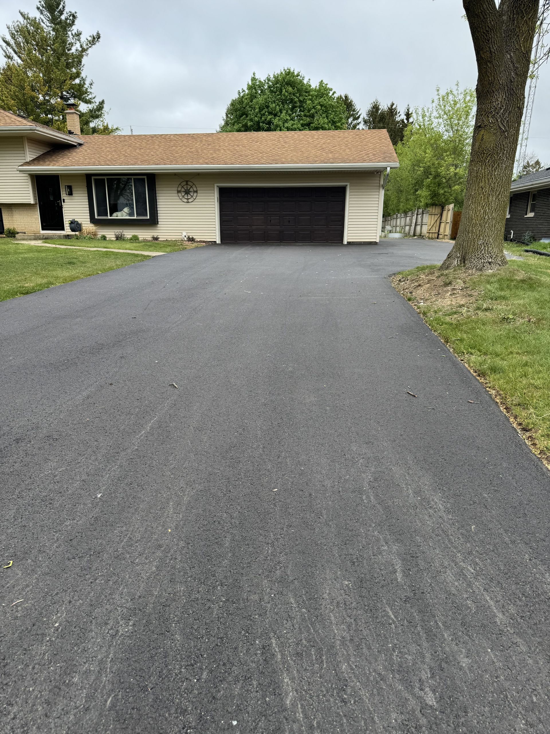 Freshly paved driveway leading to a beige house with a black garage door and large tree in front.