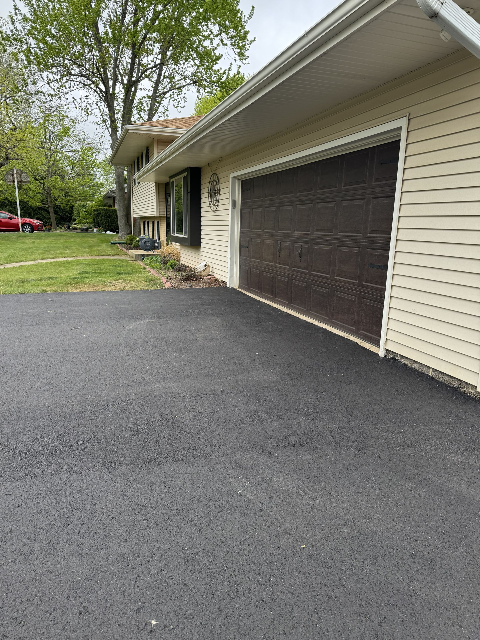 Residential driveway beside a beige house with a dark double garage door and green trees nearby