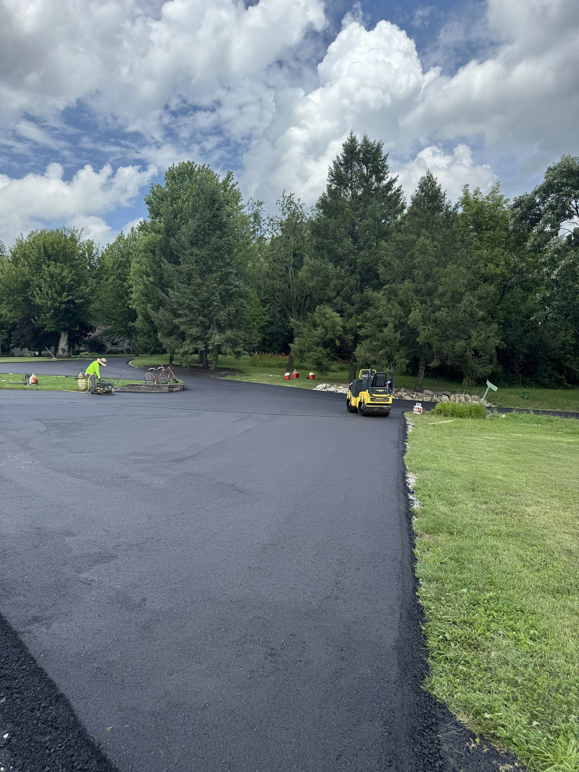 Freshly paved black driveway beside a green lawn and trees under a cloudy sky