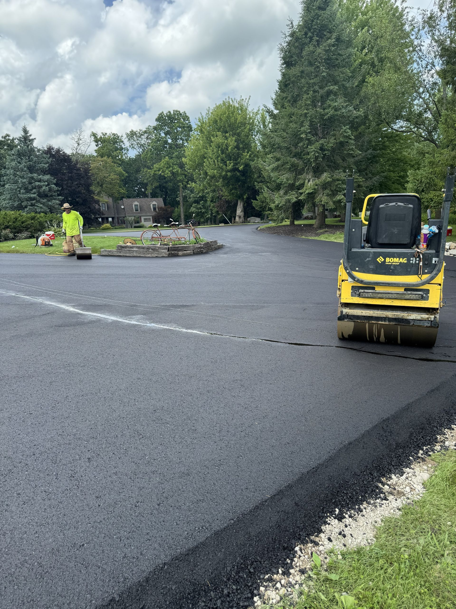 Worker operating a yellow roller on a freshly paved parking lot with trees and overcast sky nearby