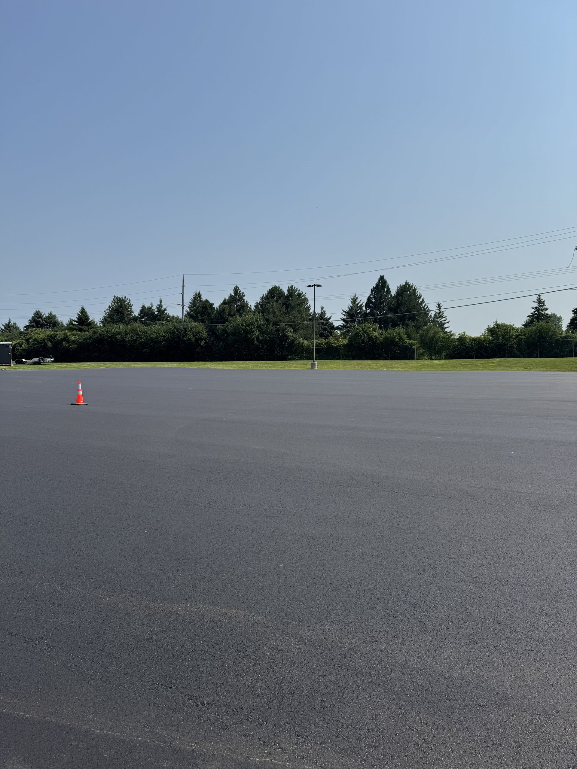 Empty paved lot with a lone orange cone, trees in the distance, and a clear blue sky