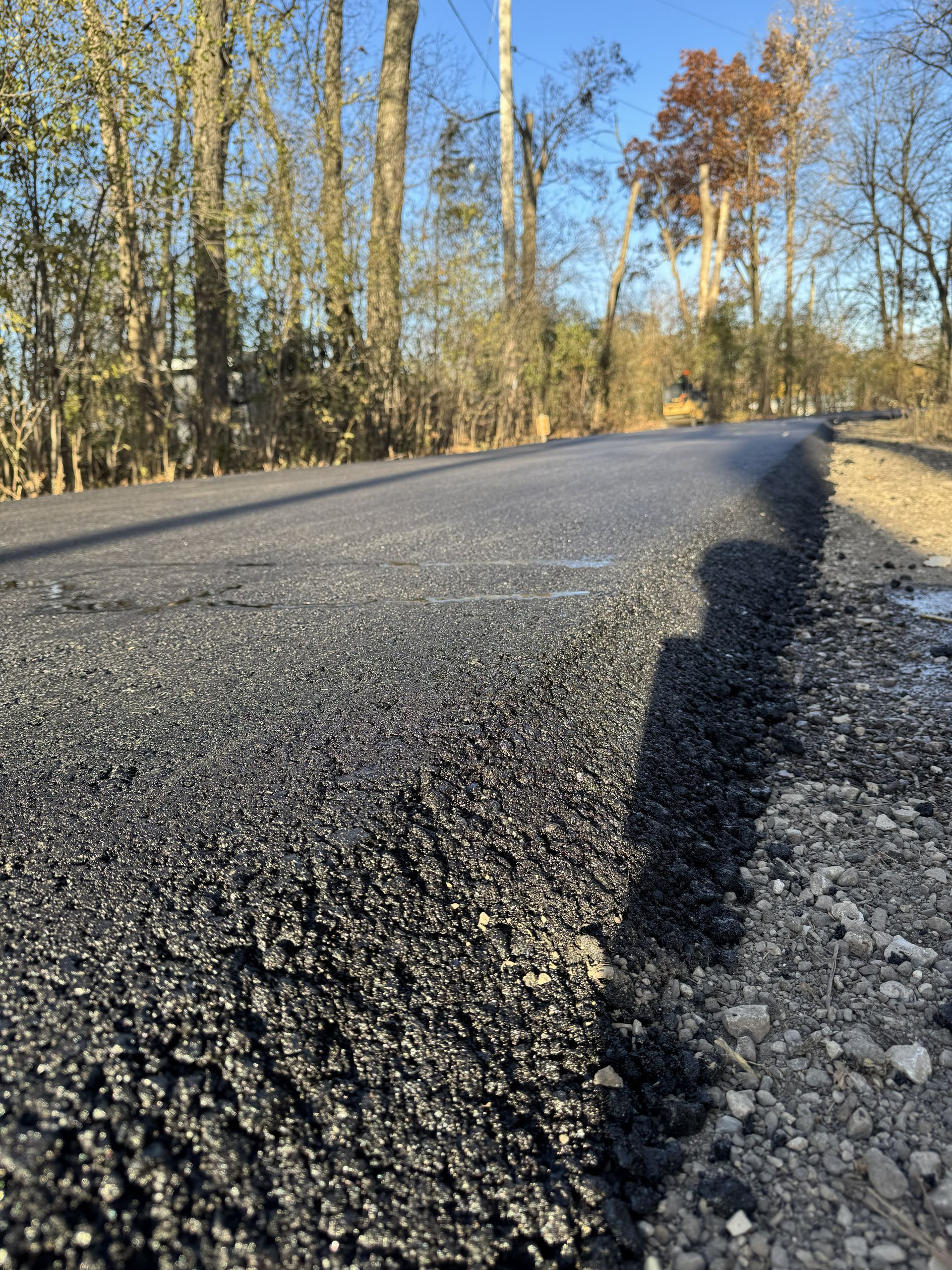 Low-angle view of a newly paved road with trees lining both sides under clear blue sky