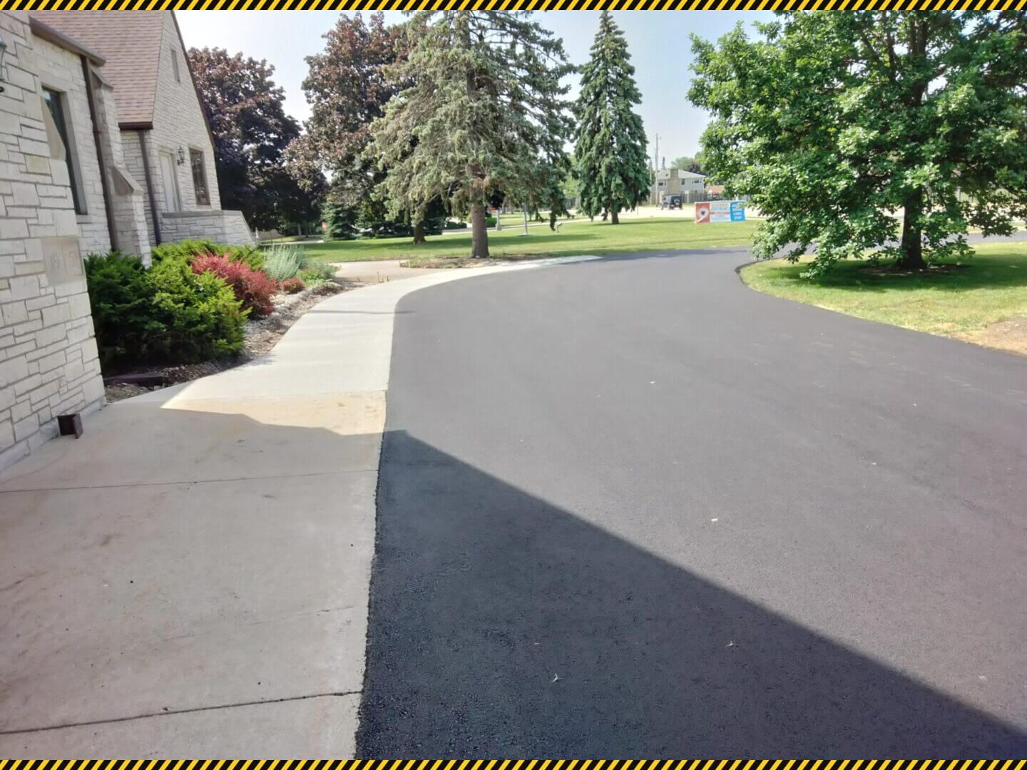 Curved paved driveway beside a stone building, with sidewalk, trees, and landscaped lawn on a sunny day
