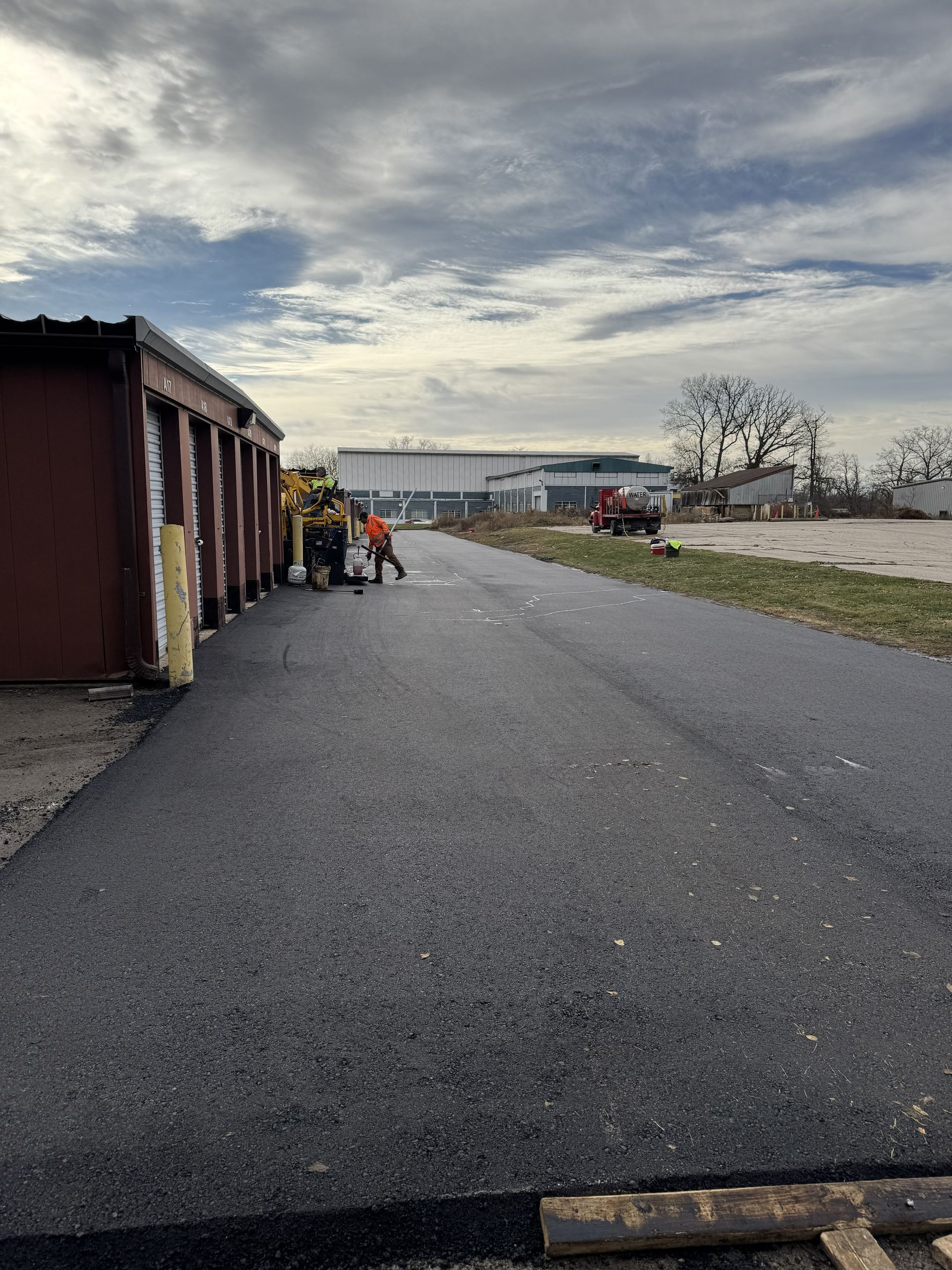 Wide paved lot beside industrial buildings under a cloudy sky, with a few people and vehicles in the distance