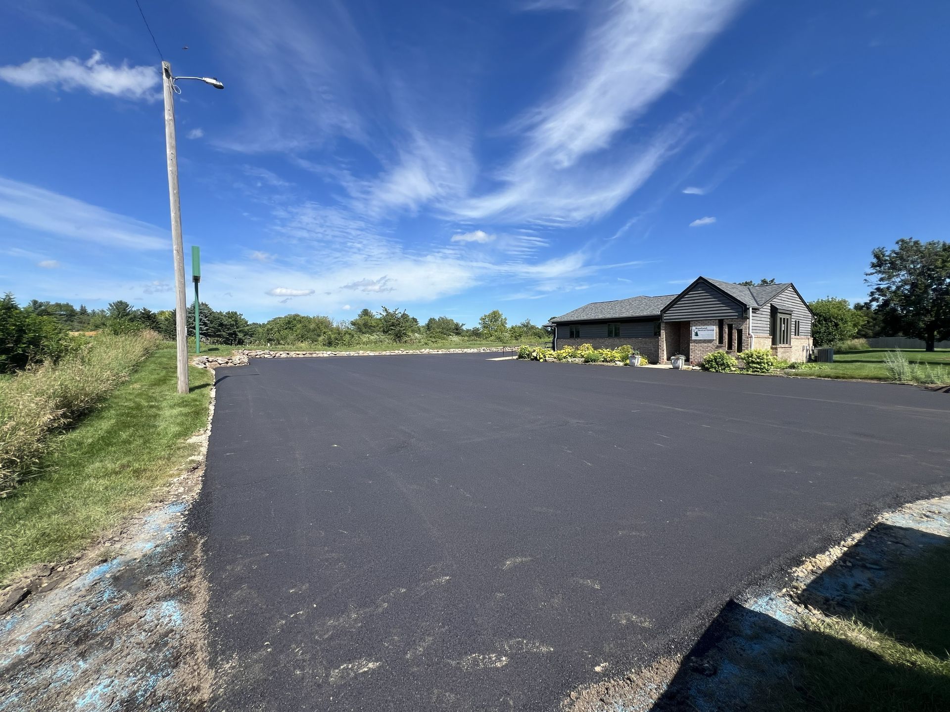 Freshly paved black road beside a small house under a blue sky with wispy clouds.