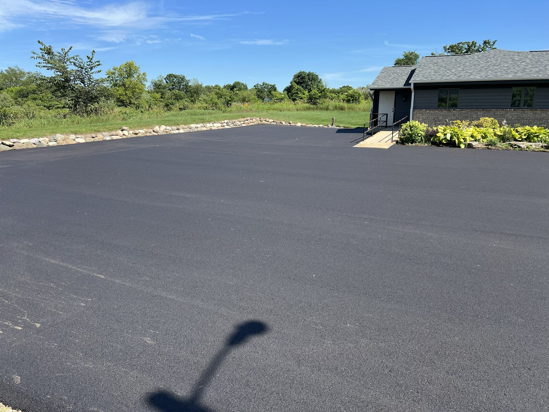 Empty blacktop driveway beside a small house and green trees under a clear sky