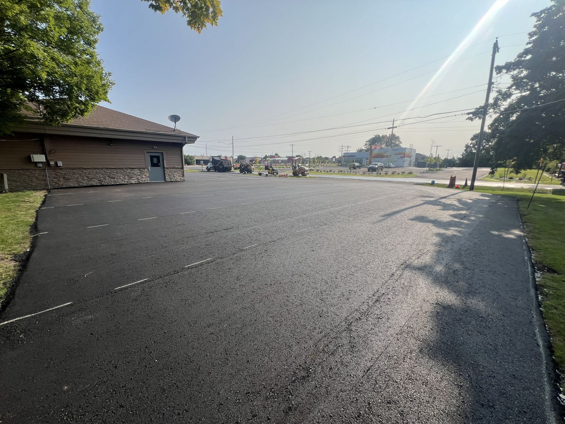 Wide asphalt lot beside a small building, with trees and bright sun in the distance.