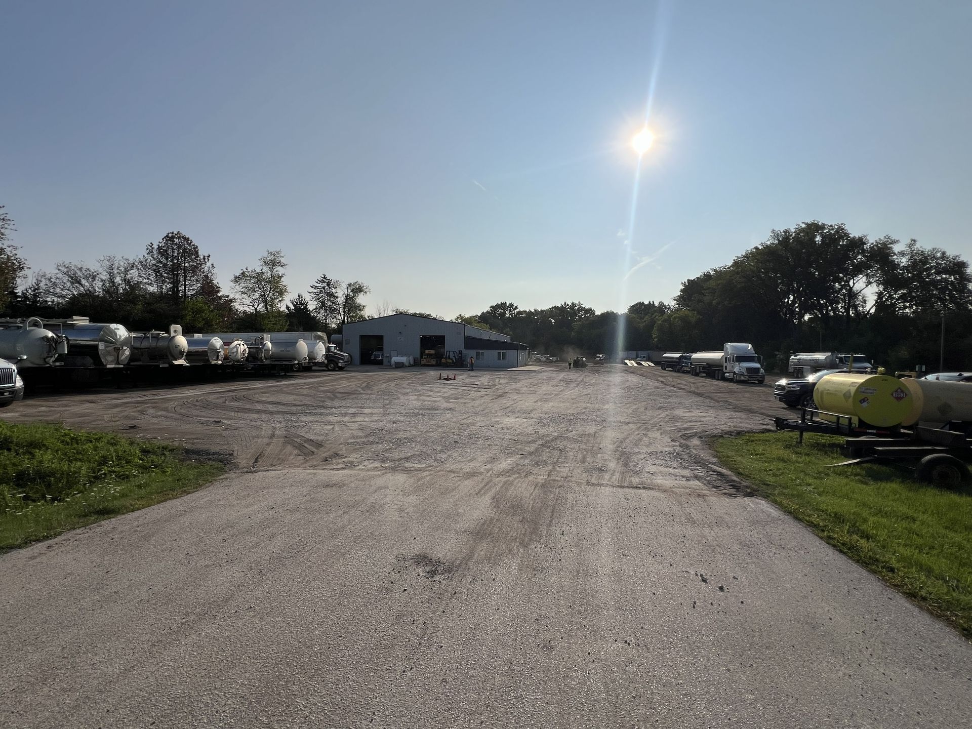 Gravel lot with parked vehicles and equipment under a bright sun, trees lining the edges.