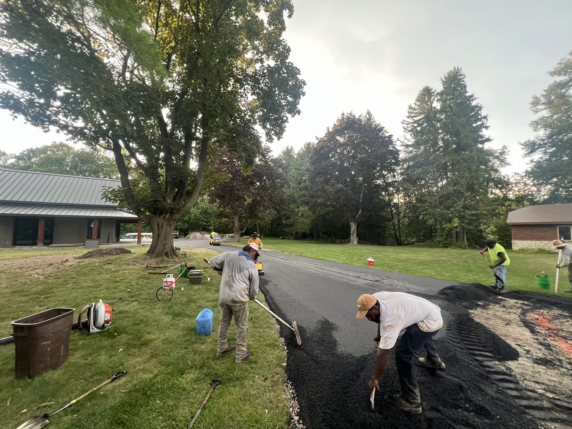 Workers paving a driveway beside a house, with gravel, tools, and trees in a yard