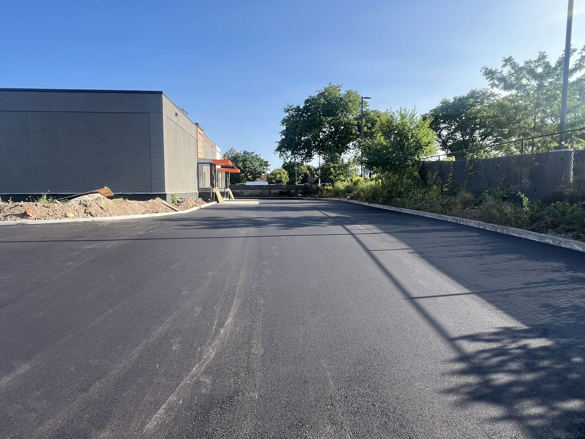 Freshly paved empty road beside a gray building and trees under a bright blue sky