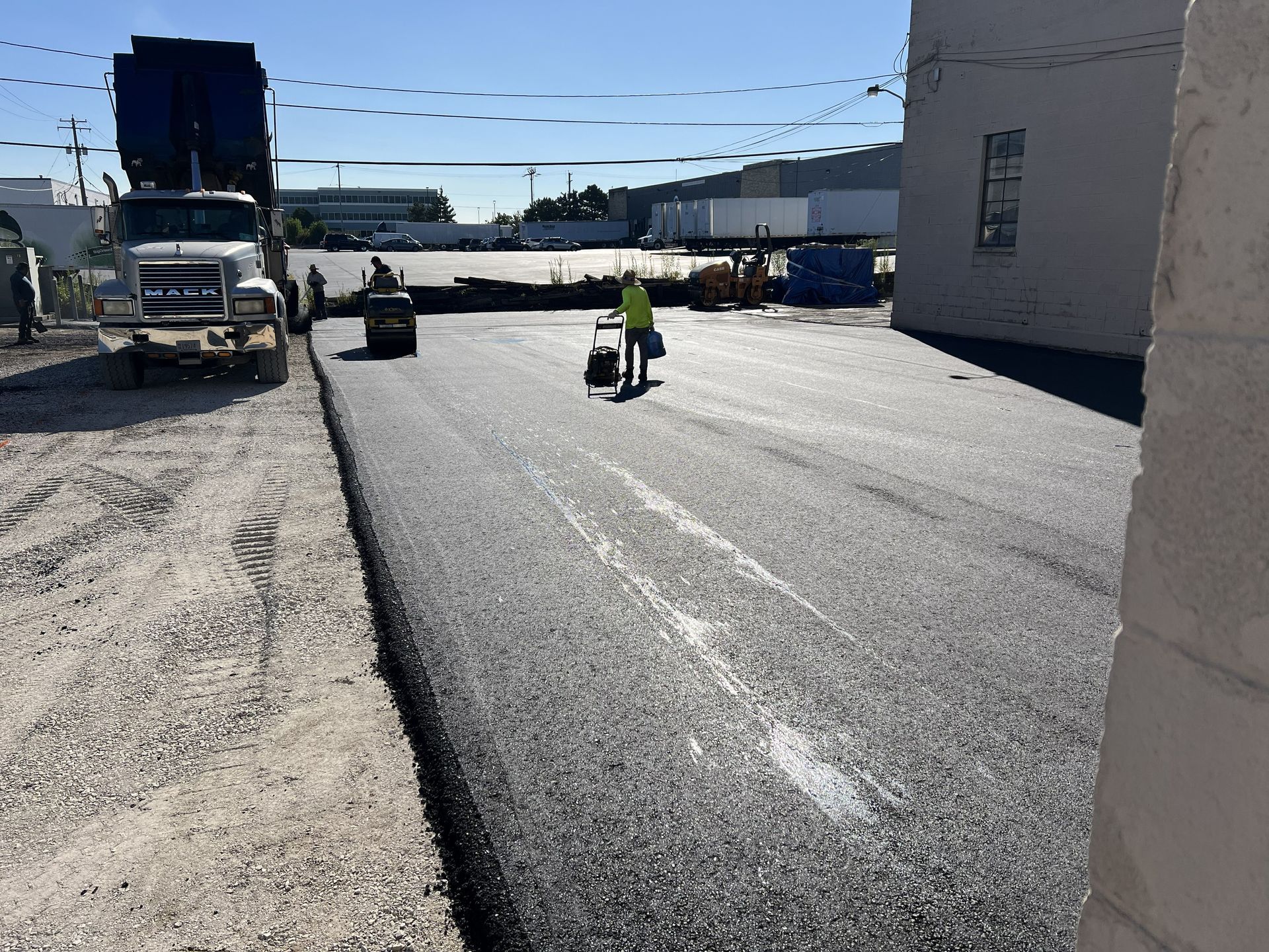 Roadwork scene with a worker in a neon vest on a paved road beside trucks and construction materials
