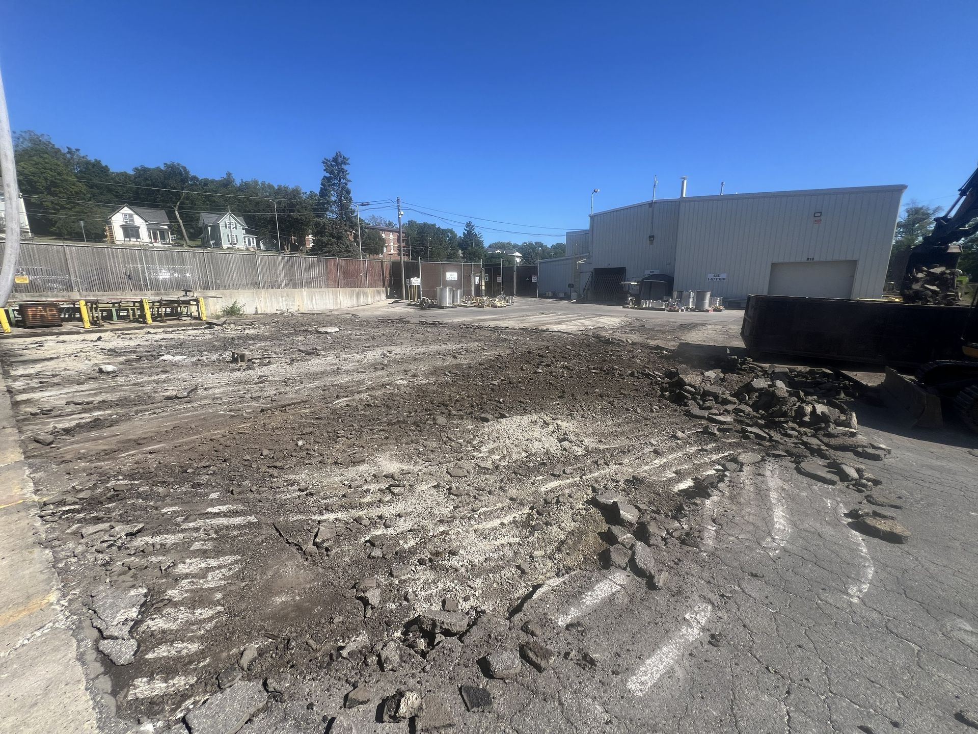 Construction site with muddy cleared ground, debris piles, and a large white building under a clear blue sky