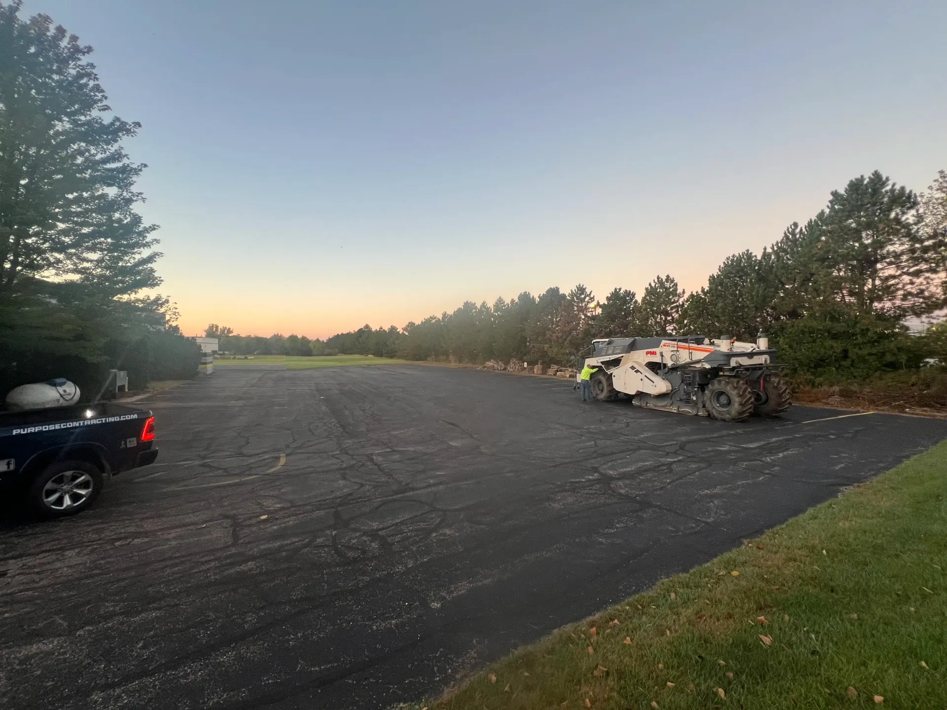 Burned-out white car wreck in a gravel lot at dusk, with trees and a black truck nearby