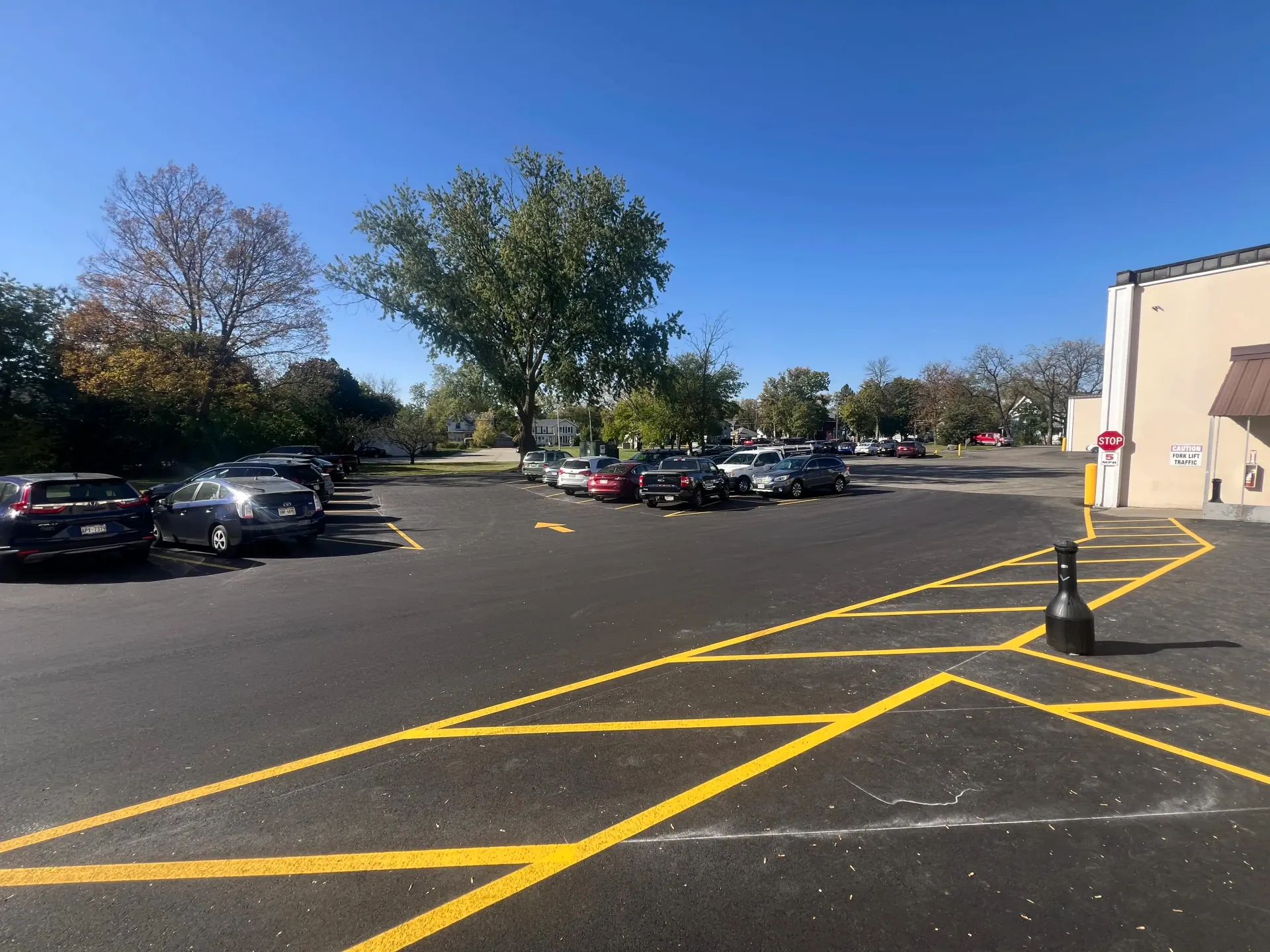 Mostly empty parking lot with parked cars, yellow crosswalk markings, trees, and a building on the right.