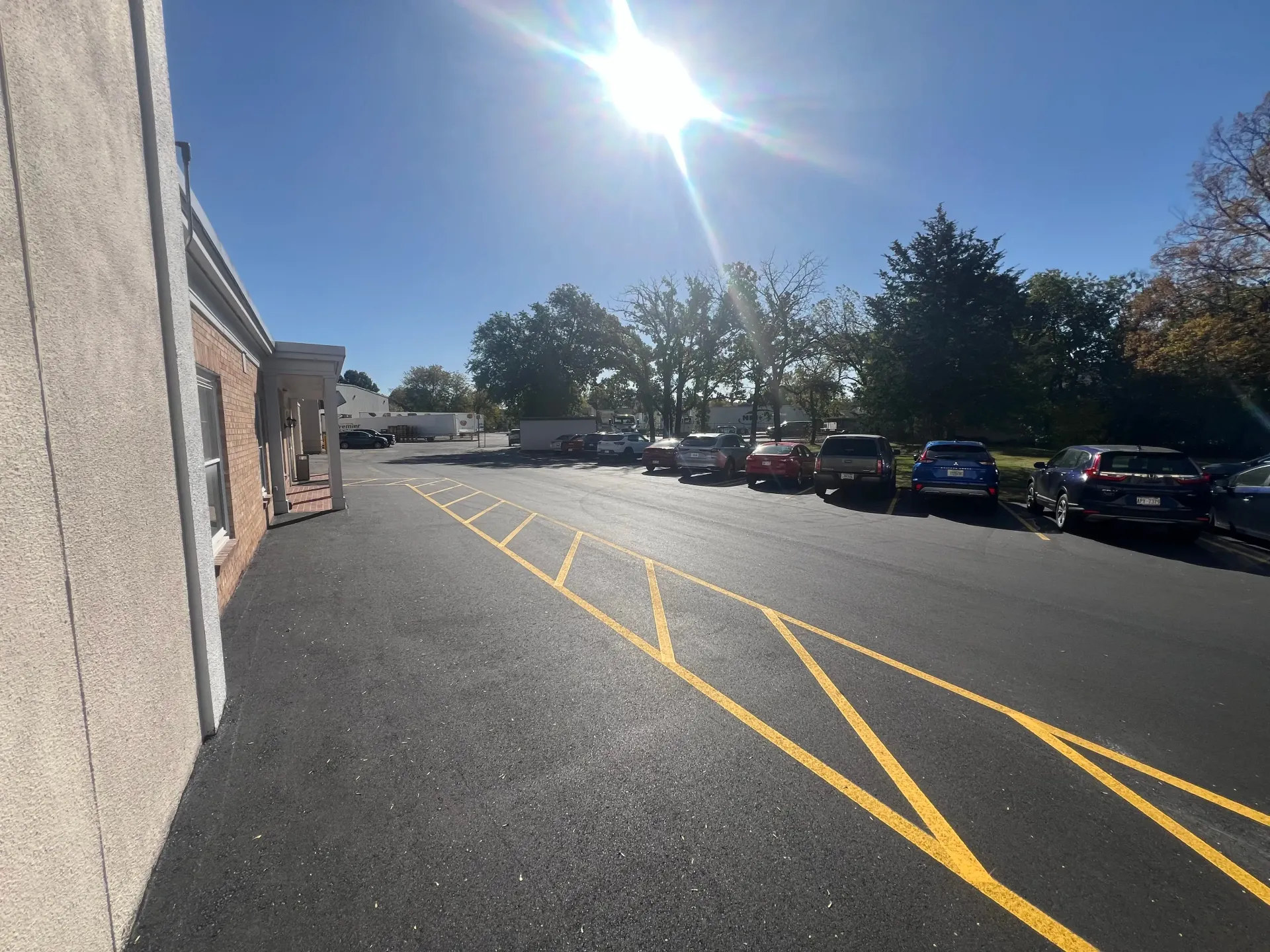 Sunlit parking lot beside a building, with parked cars and yellow marked pavement lines.