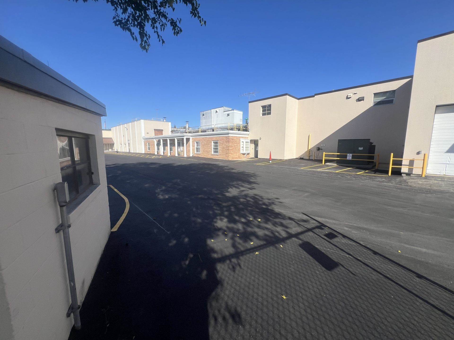 Sunlit industrial alley with beige buildings, asphalt lot, and a black dumpster under a clear blue sky