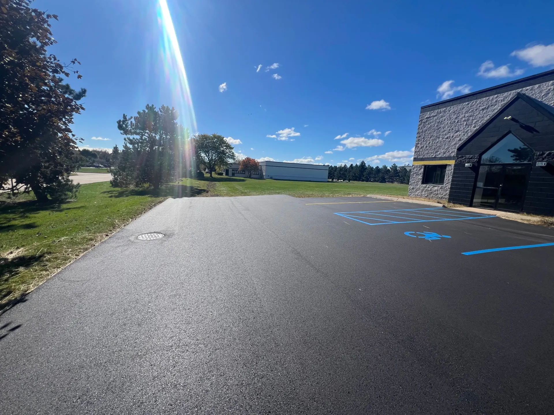 Sunlit parking lot beside a brick building, with blue handicap spaces and trees under a clear sky.
