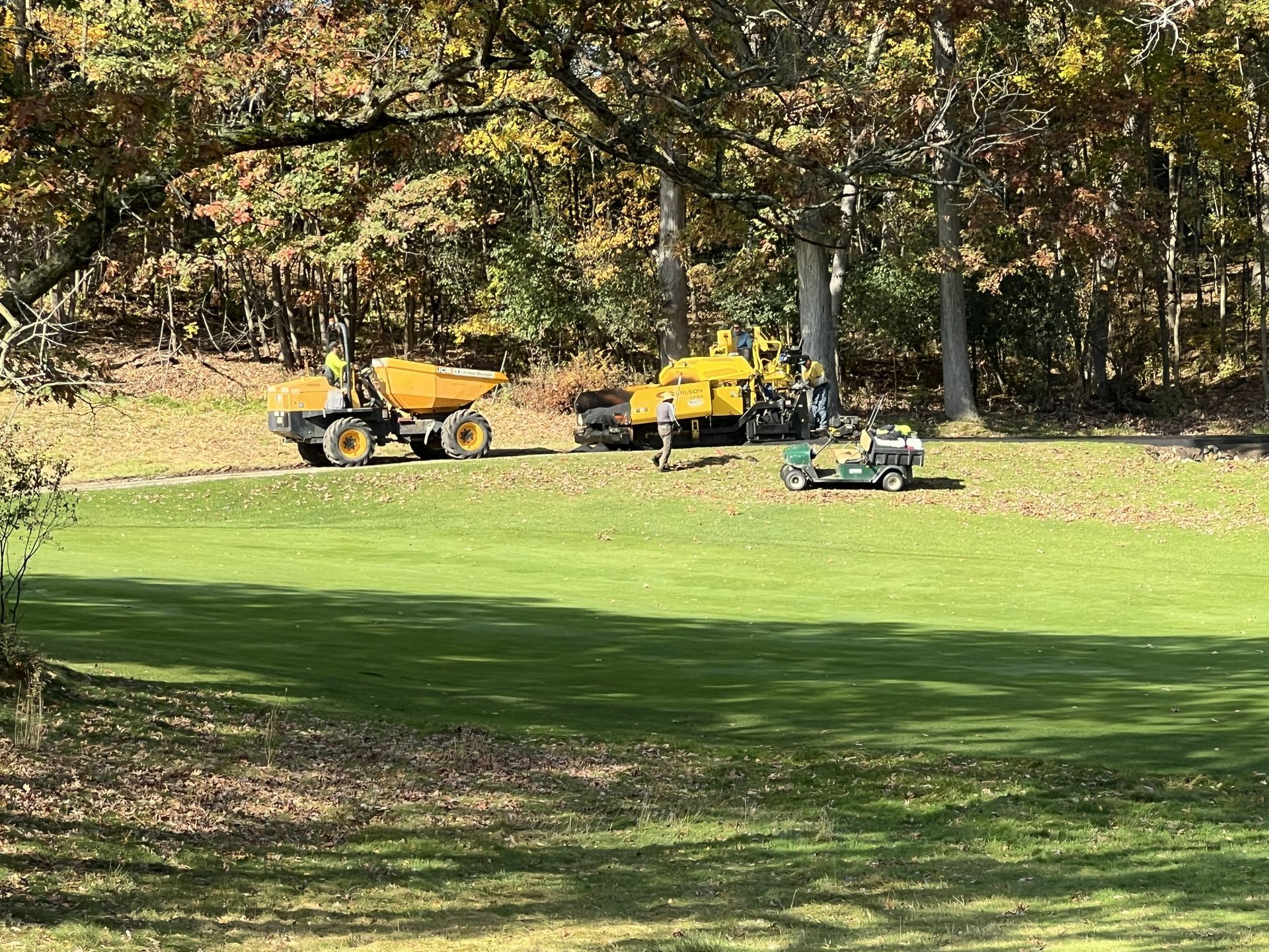 Yellow maintenance trucks and a green mower on a grassy field beside trees in autumn.
