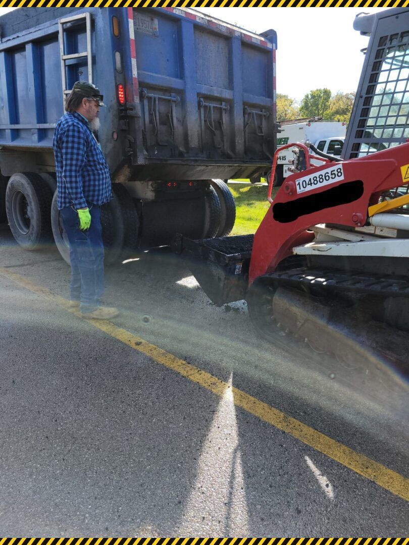 Worker stands by a dump truck and red skid steer on a road, spraying debris and water.
