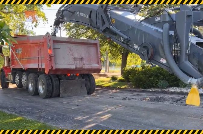 Excavator loading a red dump truck on a paved road beside trees and a yellow traffic cone