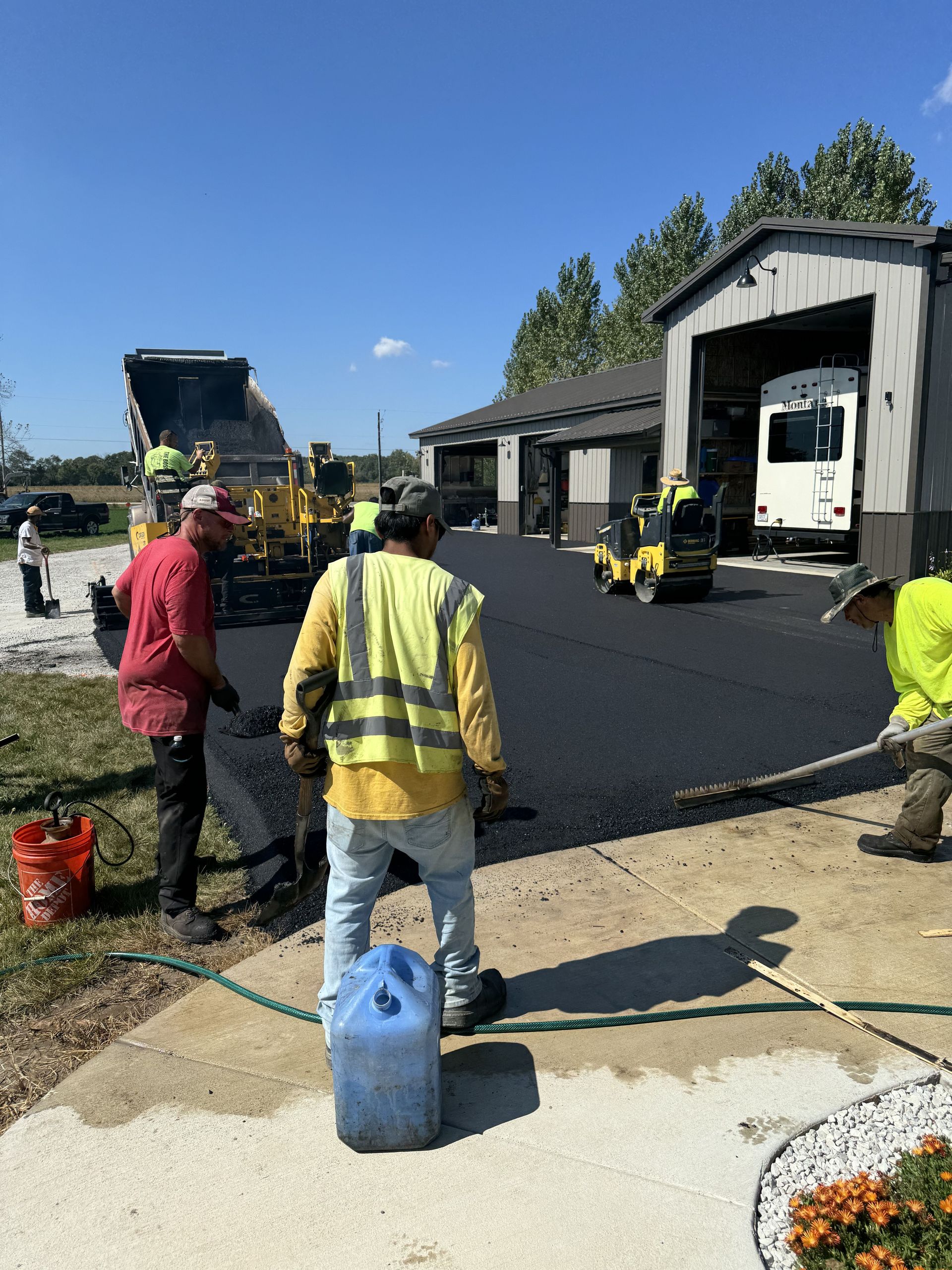 Road workers paving a lot beside a building, with a yellow loader and asphalt equipment in use.