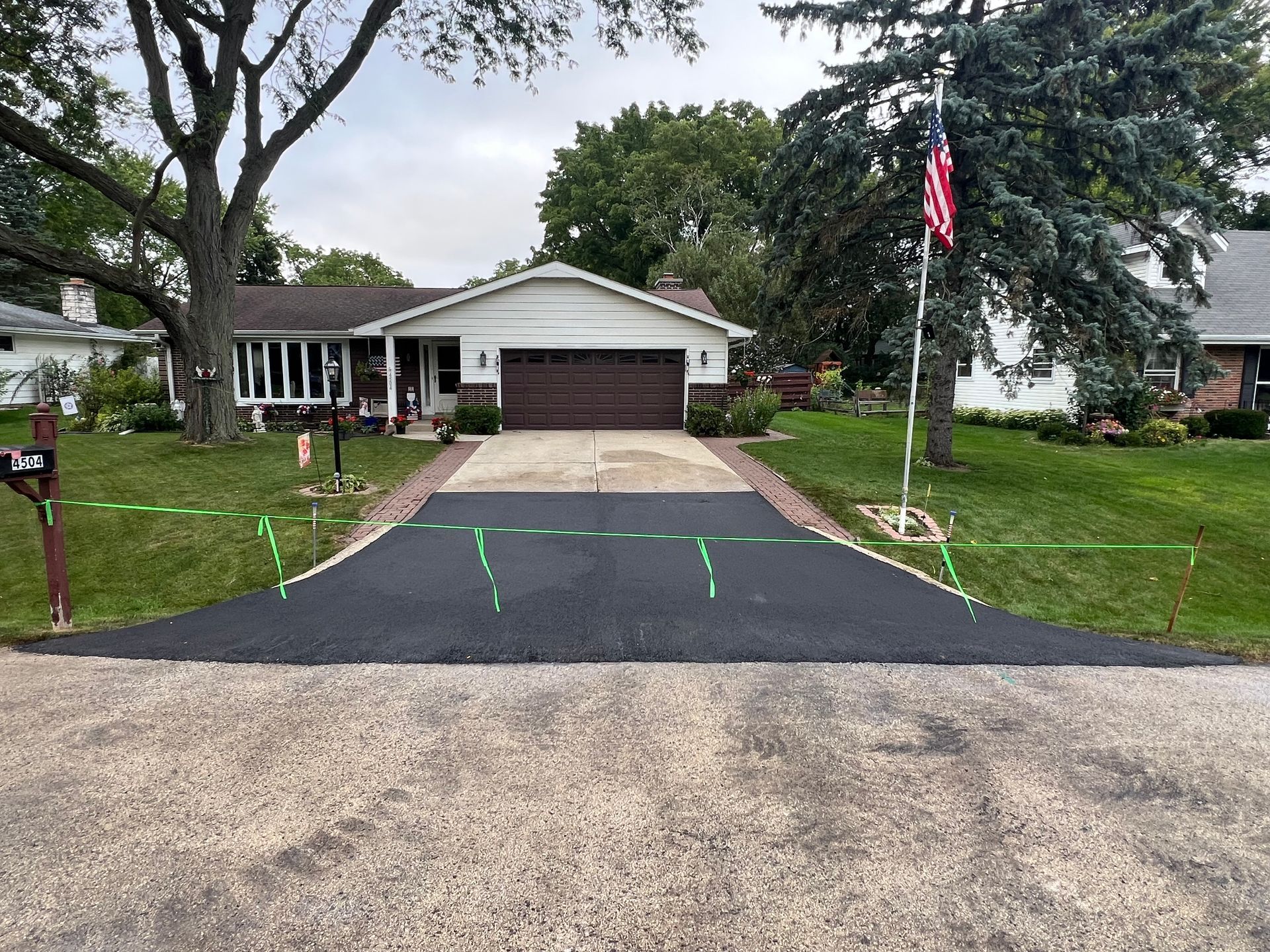 Driveway with fresh black asphalt leading to a suburban house with an American flag and trees in front.