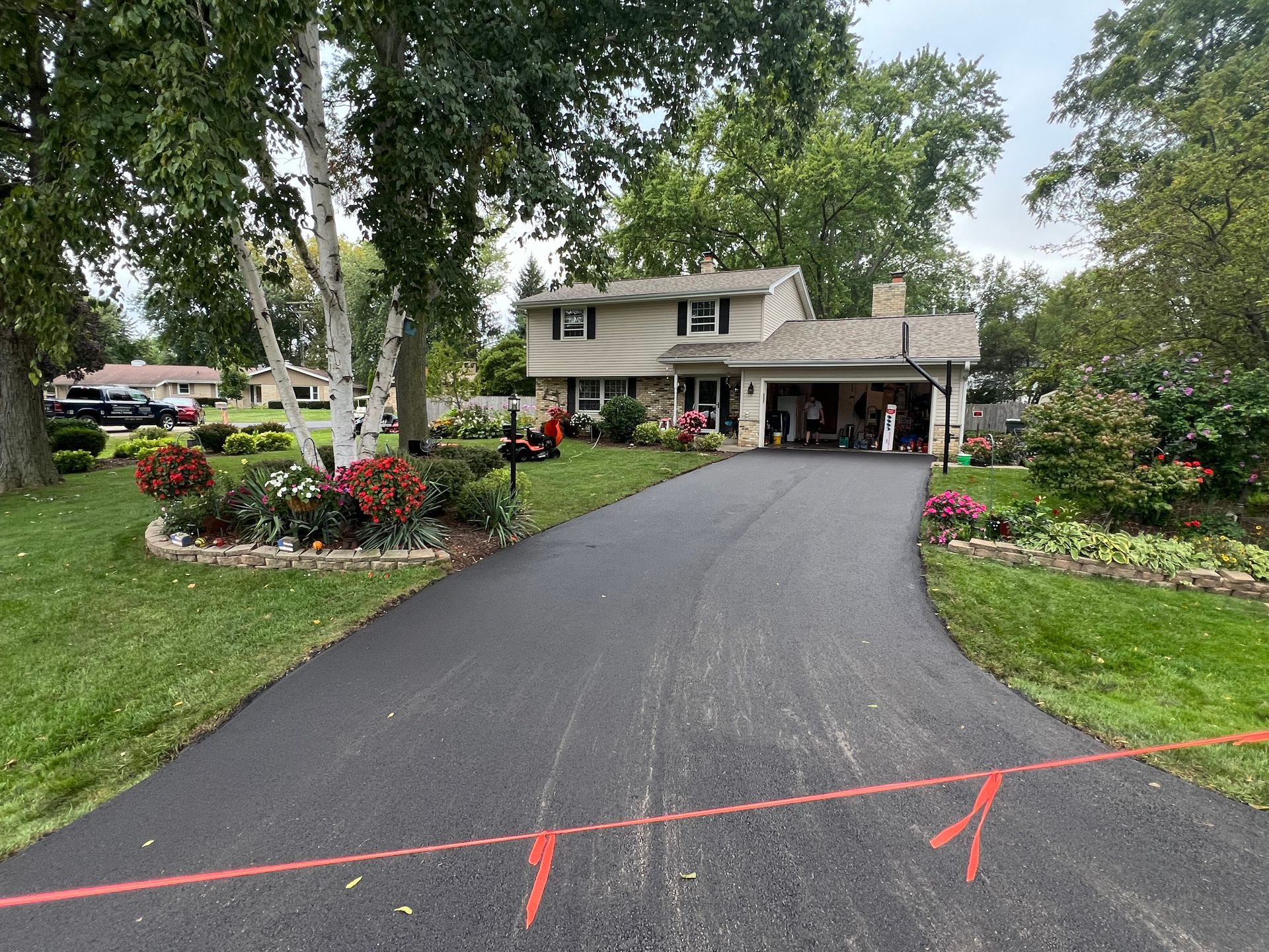 Driveway leading to a two-story house with a garage, surrounded by landscaped gardens and trees.