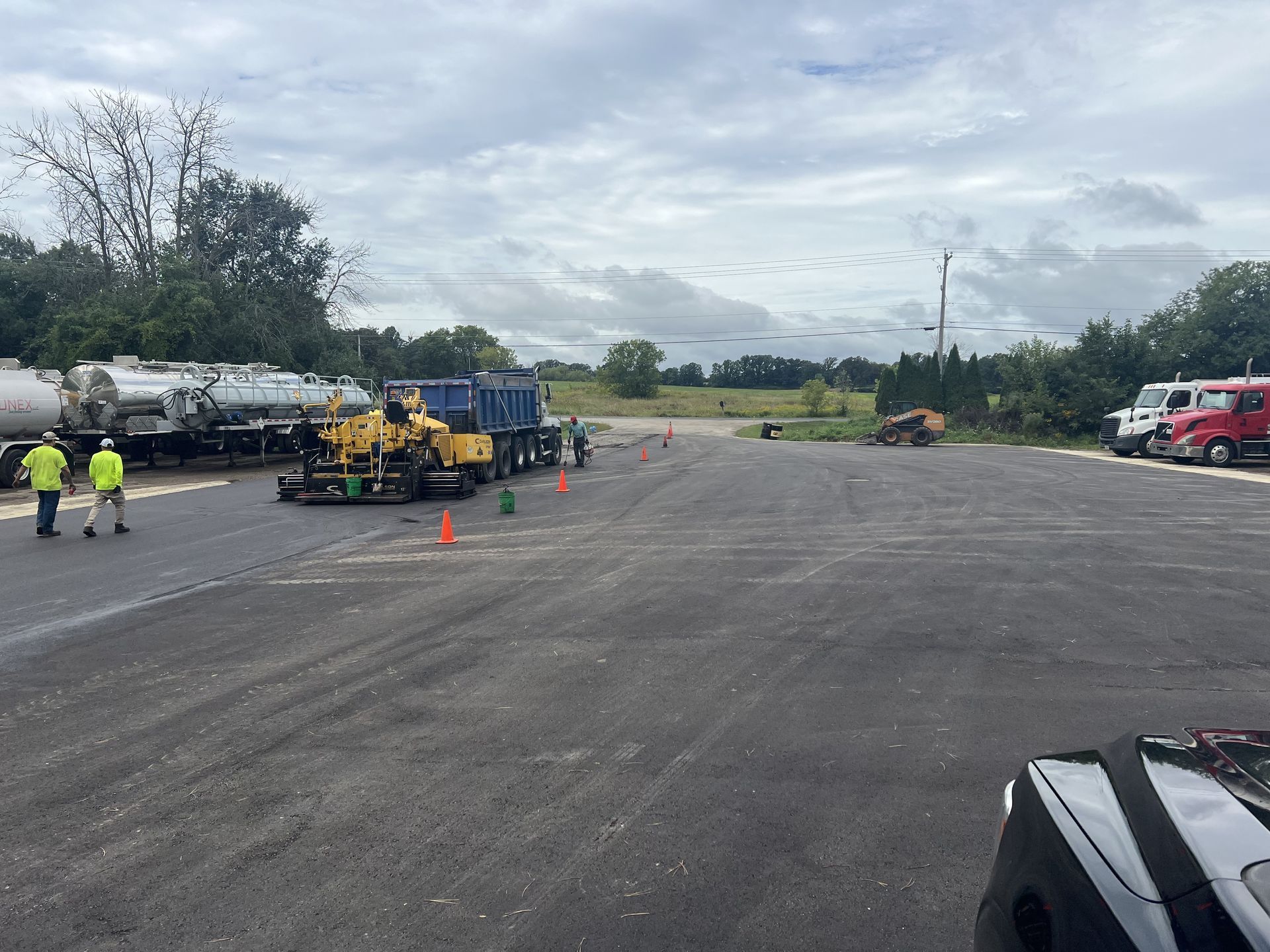 Roadwork crew and equipment on a wide paved lot with traffic cones and trucks under cloudy skies