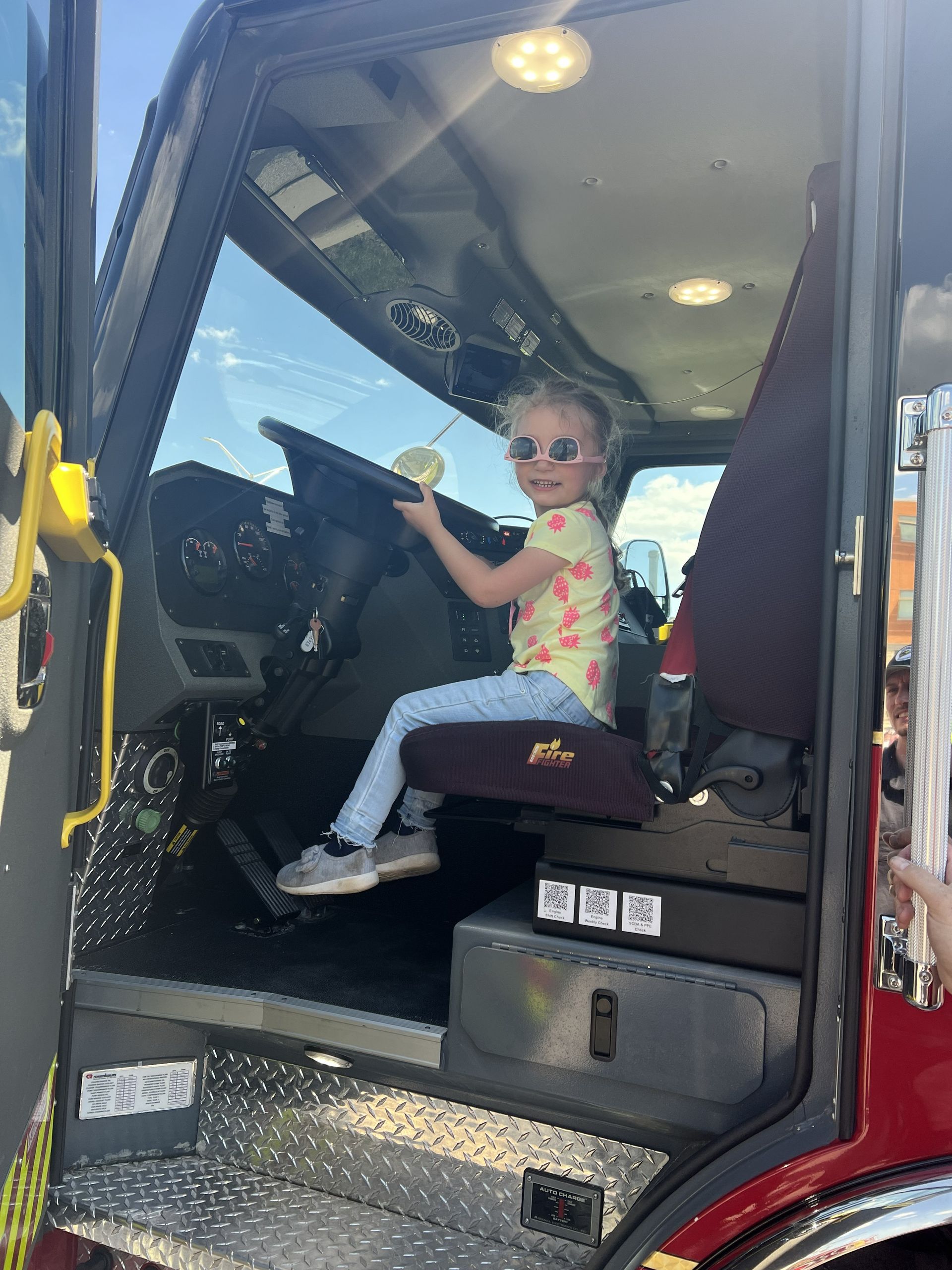 Child sitting in a fire truck cab, holding the steering wheel and wearing sunglasses.