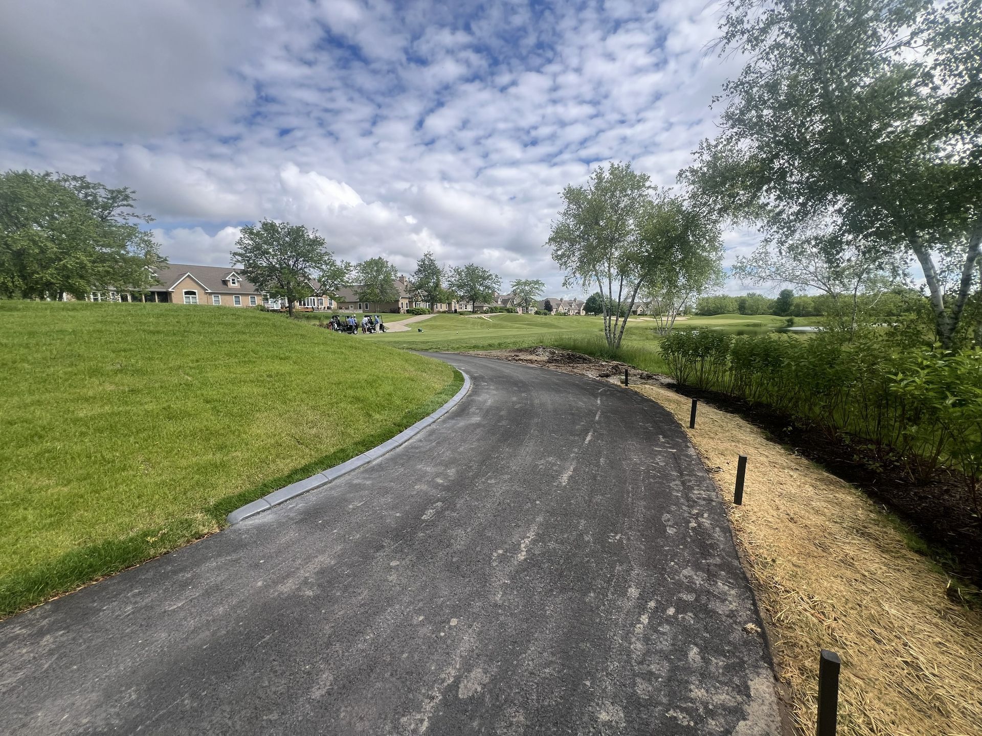 Curved asphalt path through a grassy park under a cloudy sky, with trees and shrubs lining the edges