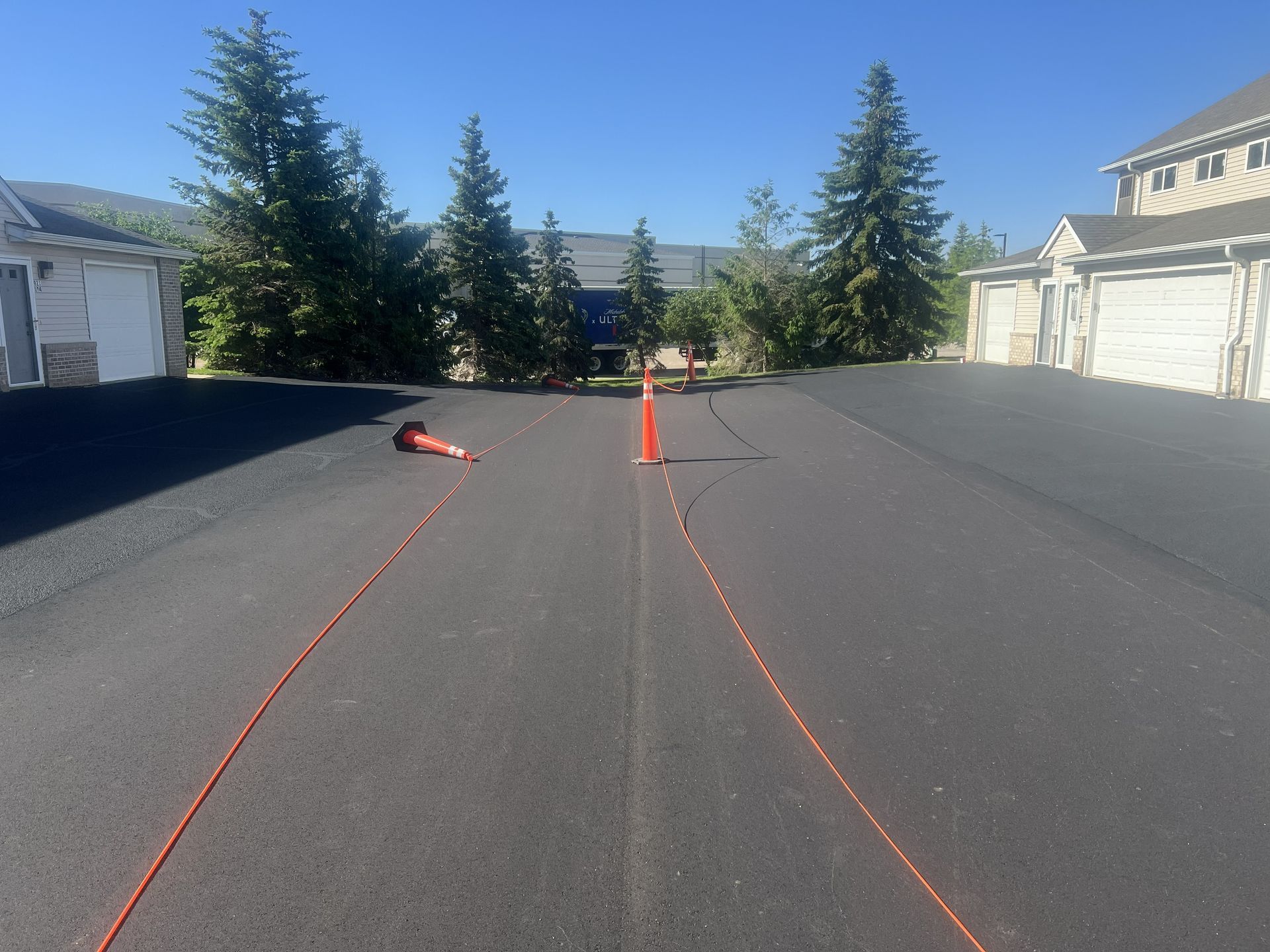 Orange cones and marker line on a paved road, with trees and houses in the background under a clear blue sky