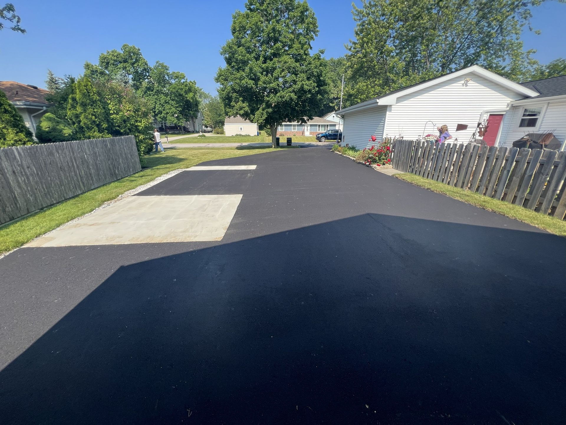 Freshly paved driveway beside a white house, with fences and trees on both sides under a sunny sky