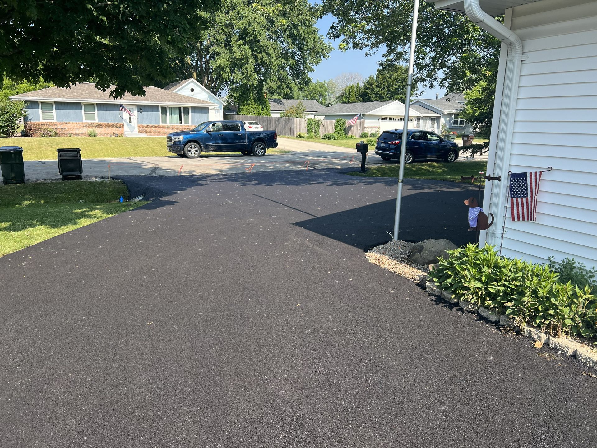 Driveway leading to a suburban street, with parked cars, a white house edge, and a small flower bed.