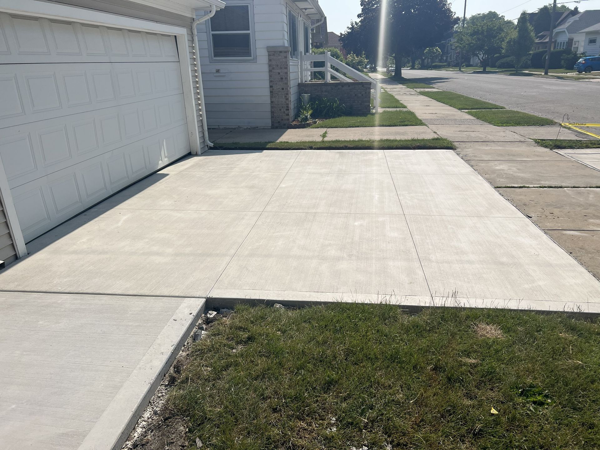 Concrete driveway beside a white garage, with a small grass patch and sidewalk on a sunny residential street