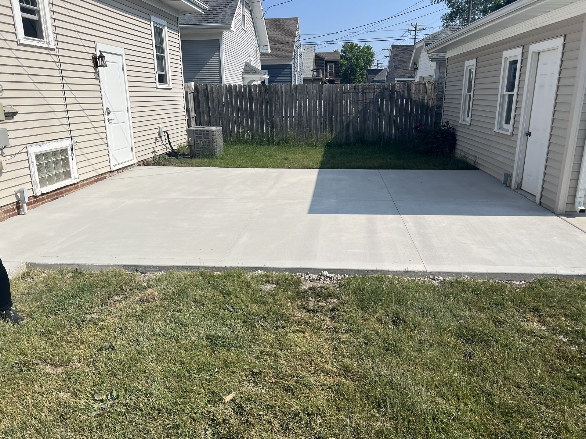 Backyard with a large concrete patio between two houses, fenced at the back, with grass in the foreground